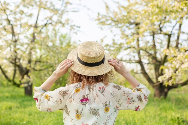 Vista posterior de una joven mujer con cabello rizado se pone un elegante sombrero de mimbre mientras camina en un jardín de flores verdes