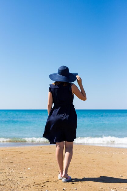 Vista posterior de la joven mujer delgada en vestido y sombrero sobre fondo de playa de verano