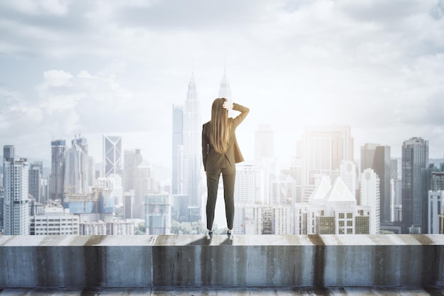 Vista posterior de una joven mujer de negocios parada en el borde de concreto de un edificio en el fondo de la ciudad con un lugar simulado en el cielo Visión futura de éxito y concepto de liderazgo