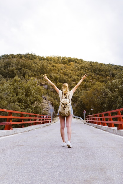 Vista posterior de la joven mujer rubia con mochila y manos arriba caminando en la carretera sobre un puente cerca de la montaña. Concepto de viaje y aventura. Viajero en medio del bosque. Viajar solo