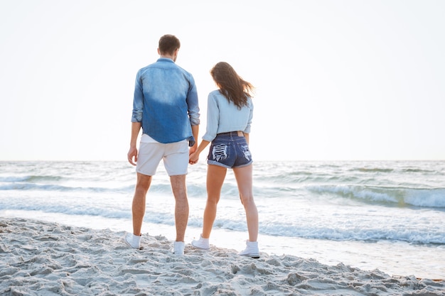 Vista posterior de una joven pareja feliz caminando y tomados de la mano en la playa.