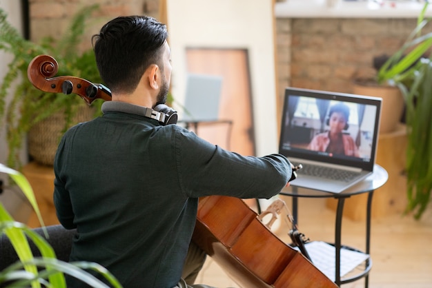 Vista posterior del joven profesor de música tocando el violonchelo frente a la computadora portátil