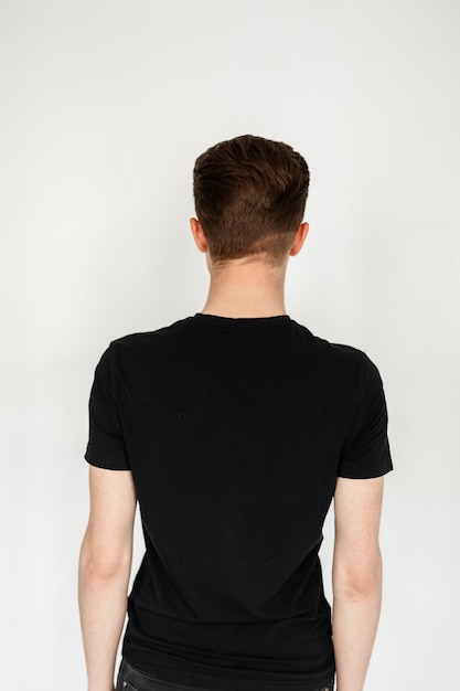 Vista posterior de joven rubio con elegante corte de pelo corto en camiseta negra de moda sobre fondo blanco en estudio. Chico elegante se encuentra frente a la pared. Broches para modelo fotográfico.