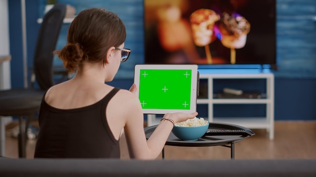 Vista posterior de una joven sosteniendo una tableta digital con pantalla verde viendo contenido de video en las redes sociales sentada en un sofá. Chica mirando un dispositivo de pantalla táctil con llave de croma mirando un vlog de influencers.