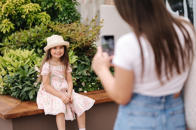 Vista posterior de mamá toma fotos de su hija en su teléfono adorable niña sentada en un banco