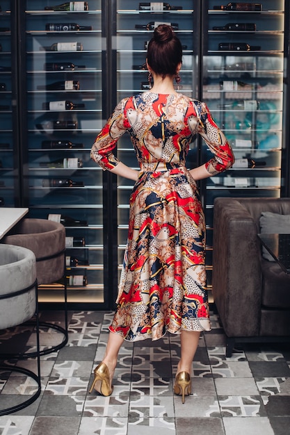 Vista posterior de una mujer adulta irreconocible con cabello oscuro en moño con vestido de seda de noche y tacones dorados sosteniendo sus manos en la cintura. Posando contra los estantes de las botellas de vino en la boutique de la bodega.