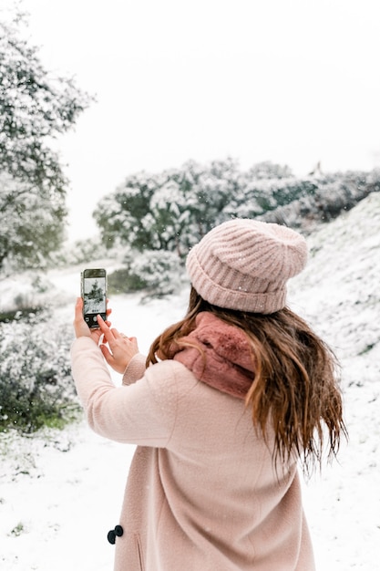 Vista posterior de una mujer anónima en ropa de abrigo abrigada de pie en el bosque y tomando fotografías del paisaje nevado mientras usa el teléfono inteligente