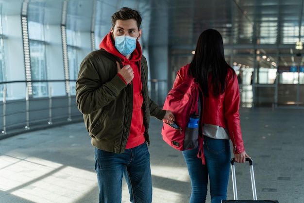 Vista posterior de la mujer con bolsa de carrito caminando por el aeropuerto mientras el ladrón usa máscara protectora robando sus cosas. Foto de stock