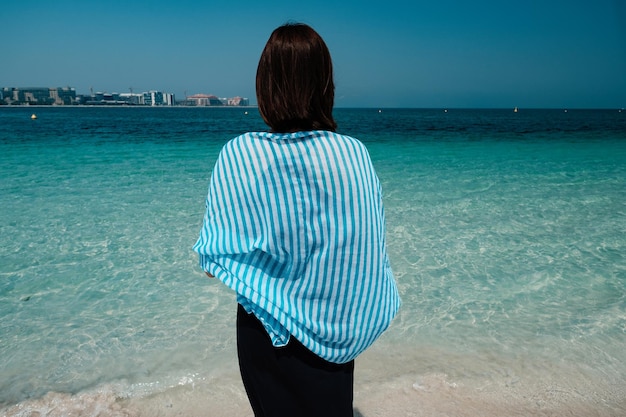 Vista posterior de una mujer caminando hacia el mar en Palm Jumeirah Dubai Una mujer con bufanda y vestido disfruta del aire salado del mar
