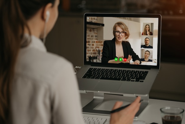 Vista posterior de una mujer en casa hablando con su jefe y otros colegas en una video llamada en una computadora portátil. La empresaria habla con sus compañeros de trabajo en una conferencia por webcam. Equipo de negocios que tiene una reunión en línea.