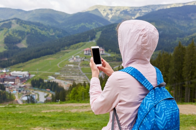 Vista posterior de una mujer delgada sosteniendo su teléfono inteligente con ambas manos, tomando fotos para el blog de viajes, buscando una nueva ruta en línea