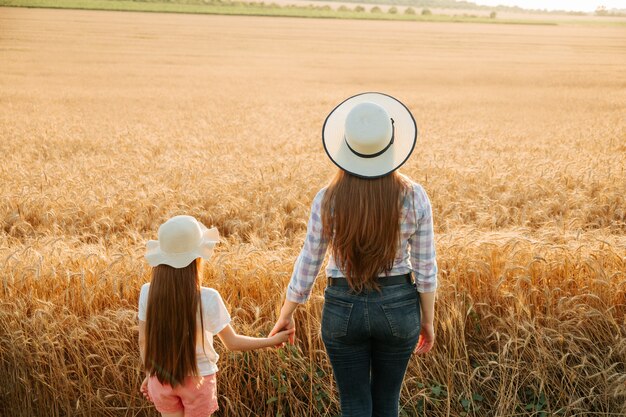 Vista posterior mujer e hija niño con sombrero en la cabeza examina tierras de cultivo trigo dorado al atardecer