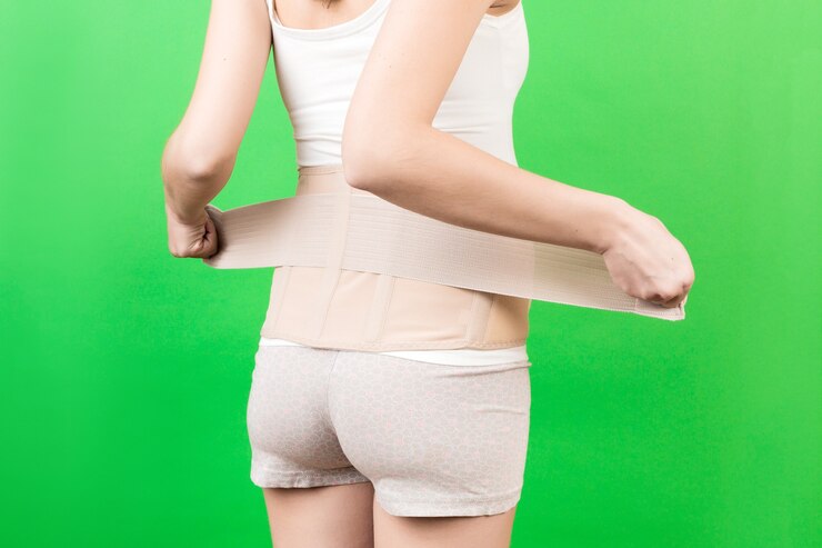 Foto vista posterior de la mujer embarazada poniéndose un vendaje de apoyo para reducir el dolor de espalda en el fondo verde con espacio de copia. imagen recortada del concepto de cinturón de soporte abdominal ortopédico.