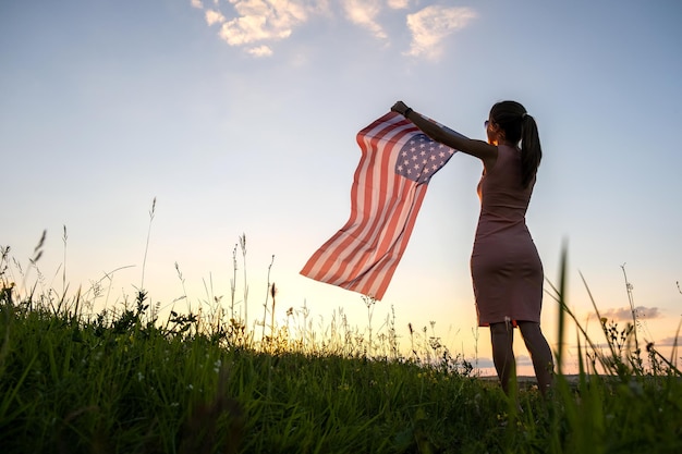 Vista posterior de una mujer feliz con la bandera nacional de EE. UU. De pie al aire libre al atardecer Mujer positiva celebrando el Día de la Independencia de los Estados Unidos Concepto del Día Internacional de la Democracia