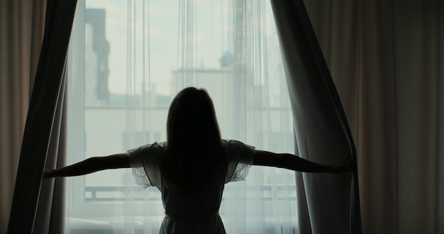 Vista posterior de la mujer joven abriendo encaje de cortina de pie en un apartamento o en un hotel moderno mirando por la ventana disfrutando de la luz del bienestar y la ciudad por la mañana