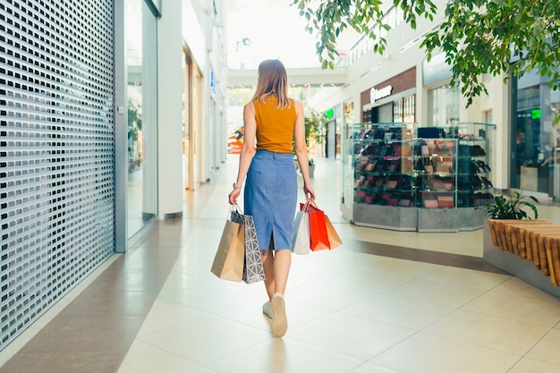 Vista posterior de una mujer joven con bolsas de regalo de colores caminando por el pasillo de un centro comercial o tienda. Compradora femenina rubia feliz satisfecha mientras hace compras en interiores. Ve y mira alrededor de la cámara