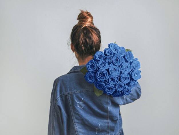 Vista posterior de una mujer joven en chaqueta de mezclilla con ramo de rosas azules clásicas en el hombro.