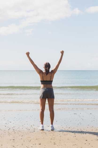 Vista posterior de la mujer joven delgada que levanta sus brazos que se colocan en la playa