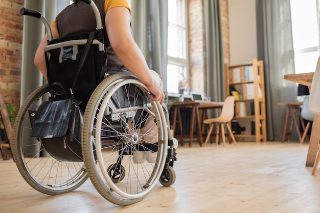 Vista posterior de una mujer joven discapacitada sentada en una silla de ruedas frente a la cámara en el centro de la habitación u oficina doméstica o moviéndose hacia la mesa
