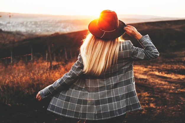 Vista posterior de la mujer joven en elegante sombrero y chaqueta de pie en el hermoso campo durante la maravillosa puesta de sol. Dama elegante irreconocible en el campo durante la puesta de sol