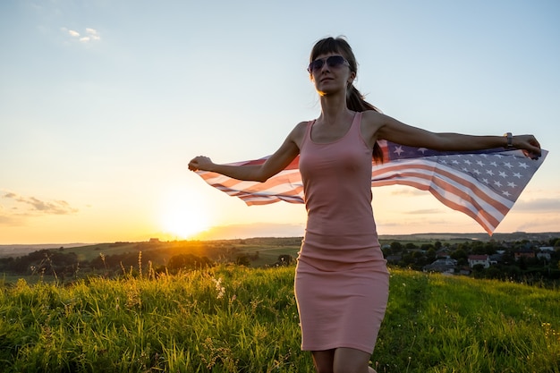 Vista posterior de la mujer joven feliz posando con la bandera nacional de Estados Unidos al aire libre al atardecer. Mujer positiva celebrando el día de la independencia de Estados Unidos. Concepto del día internacional de la democracia.