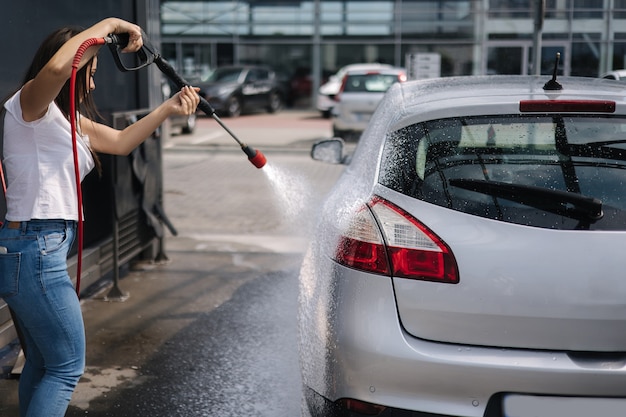 Vista posterior de la mujer joven limpiando su coche en el coche de autoservicio lavado femenino con rociador de chorro blanco