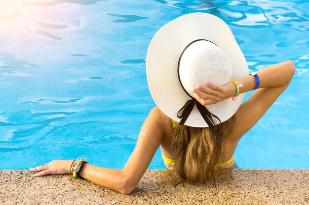 Vista posterior de la mujer joven con pelo largo con sombrero de paja amarillo relajante en la piscina de verano caliente