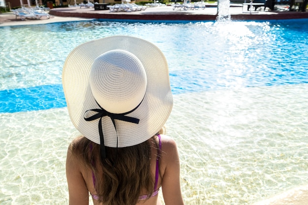 Vista posterior de una mujer joven con pelo largo con sombrero de paja relajante en verano junto a la piscina con agua azul en un día soleado.