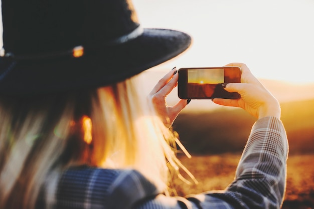 Vista posterior de la mujer joven con smartphone para tomar fotografías de la puesta de sol en un paisaje maravilloso.Mujer anónima tomando fotografías de la puesta de sol
