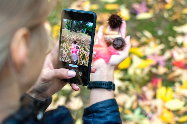 Foto vista posterior de una mujer joven sosteniendo hojas de arce naranja otoñal y conos en su mano y tomando fotografías de ellos en una cámara de teléfono inteligente en un parque de la ciudad.