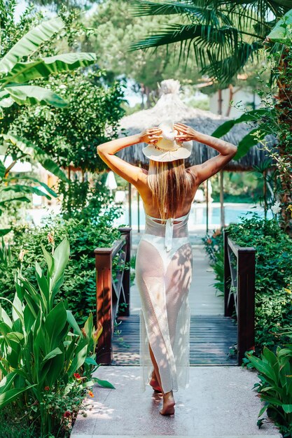 Vista posterior de una mujer joven con un vestido largo y con un sombrero caminando por el hermoso jardín tropical verde