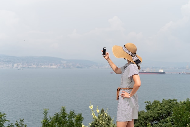 Vista posterior de la mujer joven en vestido de verano y sombrero tomando fotografías del paisaje urbano mediante teléfono móvil