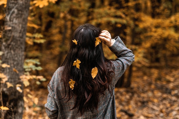 Vista posterior de la mujer morena con hojas amarillas de otoño en el pelo largo sobre fondo de naturaleza otoñal