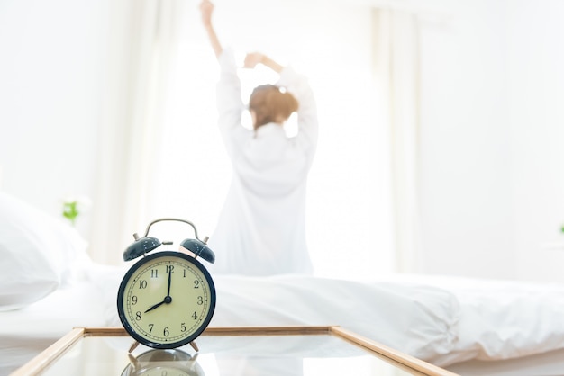 Vista posterior de la mujer que se extiende en la mañana después de despertarse en la cama junto a la ventana con reloj despertador