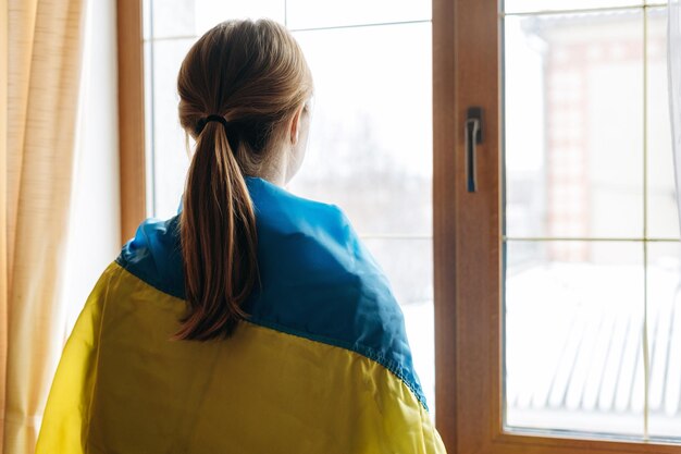 Vista posterior de la mujer rubia envuelta en la bandera de Ucrania mirando por la ventana Apoyo al concepto de Ucrania