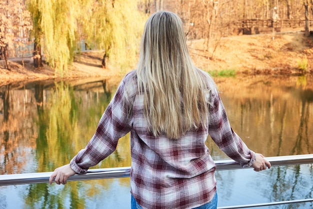 Vista posterior de una mujer rubia junto al lago con el telón de fondo de un hermoso paisaje otoñal