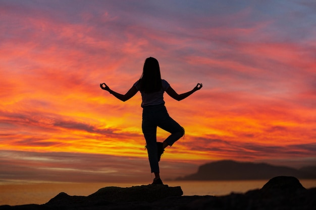 Vista posterior de una mujer satisfecha de felicidad tranquila y libre parada en la roca superior con posición de yoga contra el idílico cielo de la puesta de sol junto al mar en un feliz y hermoso momento inspirado de su vida