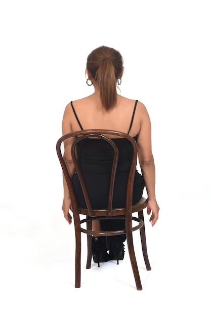 Vista posterior de una mujer sentada en una silla con vestido y zapatos de tacón sobre fondo blanco.