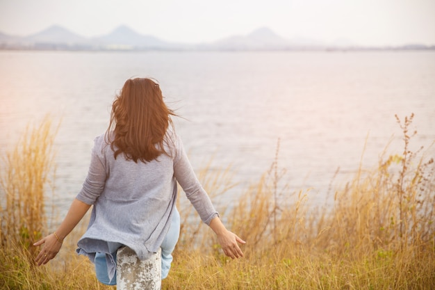 La vista posterior de la mujer se sienta al lado de la carretera con el árbol alrededor, el soporte asiático de la muchacha del viajero da vuelta atrás y mira lejos