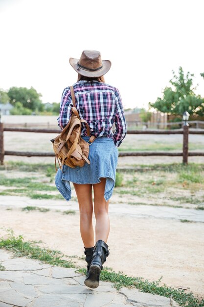 Vista posterior de la mujer vaquera con sombrero con mochila caminando en el rancho