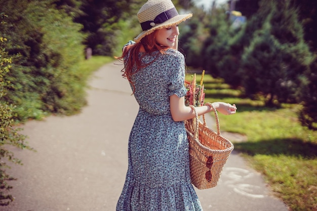 Vista posterior de la niña pelirroja adulta con elegante bolso de paja y sombrero de paja que camina en el parque al aire libre