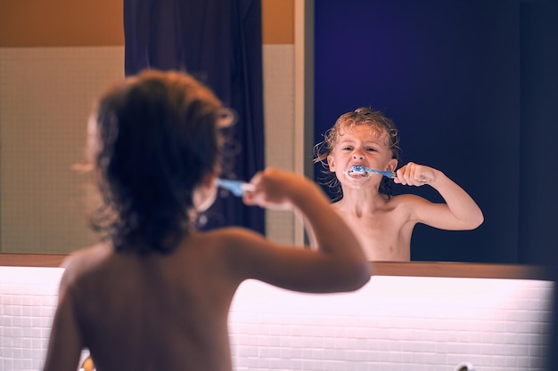 Vista posterior de un niño sin camisa con el pelo mojado reflejándose en el espejo y cepillándose los dientes durante la rutina matutina en el baño