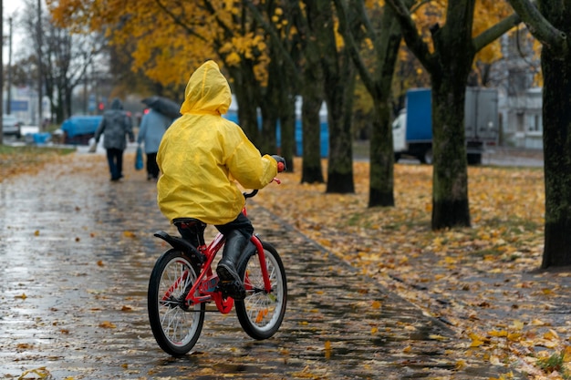 Vista posterior del niño en impermeable amarillo. Niño monta en bicicleta en un parque de otoño bajo la lluvia.