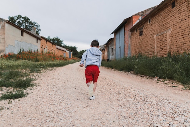 Foto vista posterior de un niño irreconocible en pantalones cortos rojos y una sudadera gris corriendo al aire libre en un casco antiguo