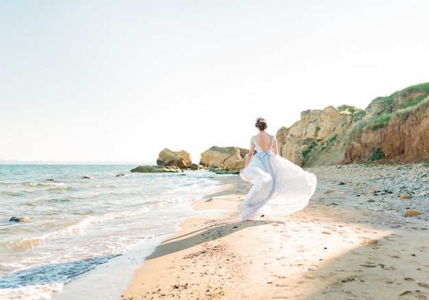 Vista posterior de la novia bonita morena con vestido de novia de gasa ligero bordado con cuentas corre cerca del mar.