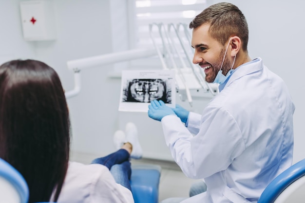 Vista posterior de una paciente discutiendo con un dentista masculino mirando rayos en una clínica moderna