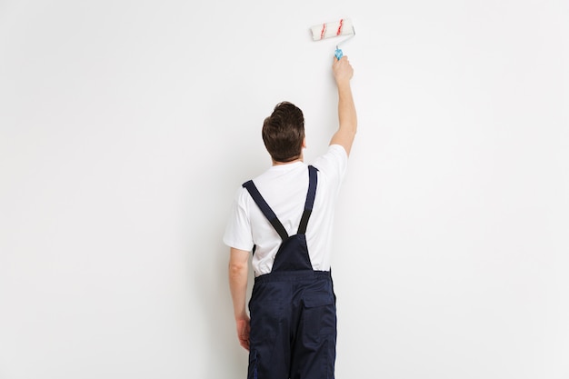 Vista posterior de la pared de pintura joven constructor masculino con rollo de pintura sobre pared gris