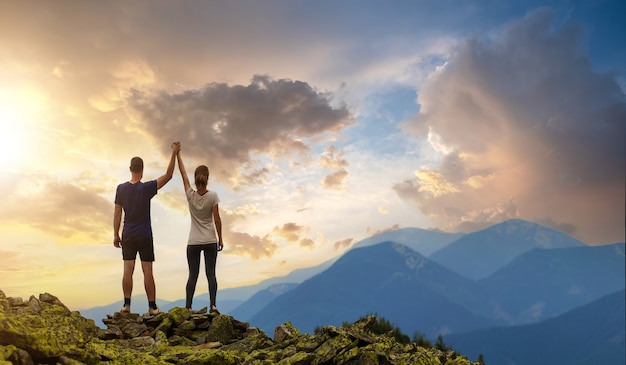 Vista posterior de la pareja joven excursionista de pie con los brazos levantados cogidos de la mano en la cima de la montaña rocosa disfrutando del panorama del atardecer.