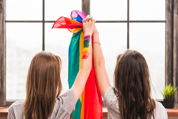 Vista posterior de una pareja de lesbianas sosteniendo las manos y la bandera del arco iris mirando la ventana