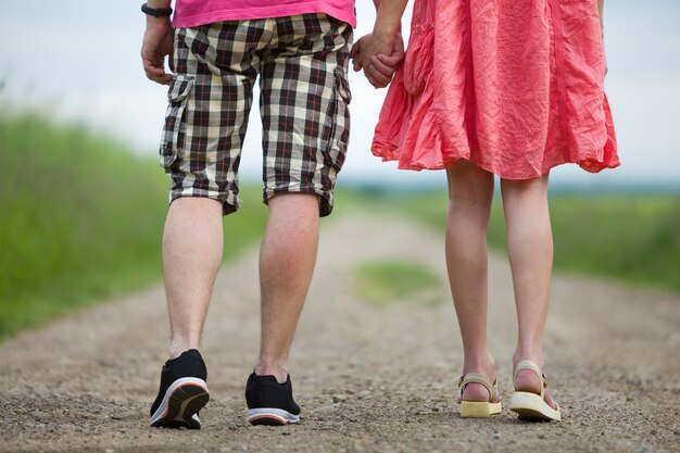 Vista posterior de las piernas de la joven mujer delgada en vestido rojo y hombre en pantalones cortos caminando juntos por camino de tierra en un día soleado de verano en escena borrosa.